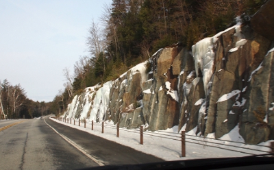 [Chunks of rock on the tree-topped hillside with frozen falls of ice among the rock on the right side of the road.]